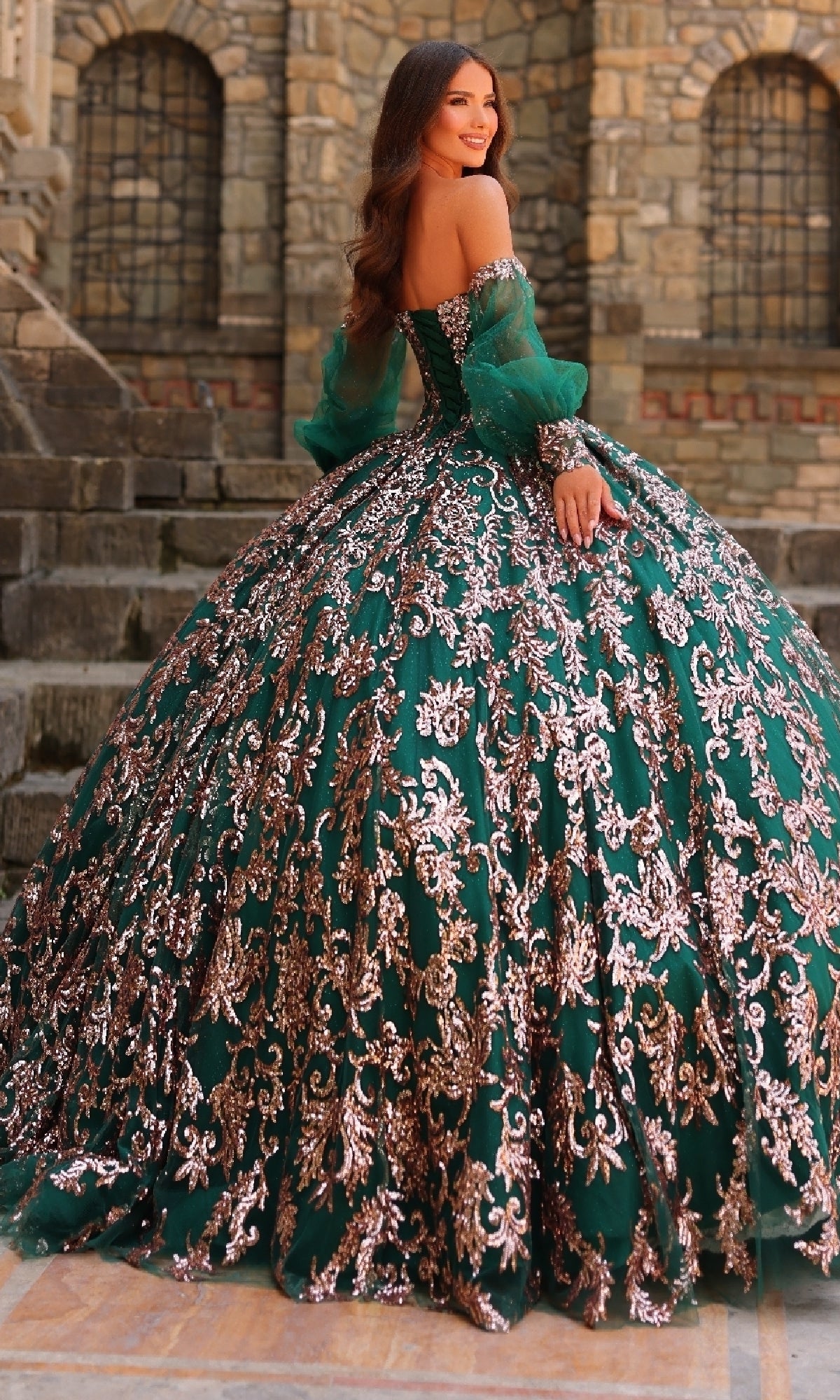 A woman stands on stone steps wearing the Amarra 54238 Sequin Quinceanera Dress, featuring a corset bodice, silver embroidery, and a voluminous green skirt.